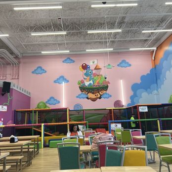 Kids Empire Paramus - Tables/Seating. Infant/toddler area on that back wall - Children's indoor play area with a mural of a cartoon creature on the wall, surrounded by clouds and colorful decorations, with seating and tables in the foreground. - Kids Activities Near Me - Paramus, New Jersey