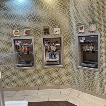 Yofruito - February 2022 - Three ice cream vending machines are installed in a wall covered with small, multicolored mosaic tiles. Each machine has a digital control panel and is labeled with flavor options such as mango, watermelon, chocolate, vanilla, strawberry, and original. The area is illuminated by ceiling lights, and the floor features a combination of beige and dark brown tiles. - Ice Cream & Frozen Yogurt Near Me - Buffalo Grove, Illinois