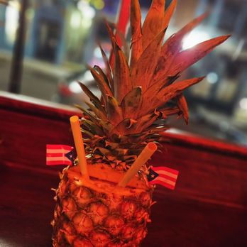 Papi - A pineapple with two yellow straws and small American flags inserted into it, placed on a dark surface with a blurred background. - Cocktail Bars Near Me - Portland, Maine