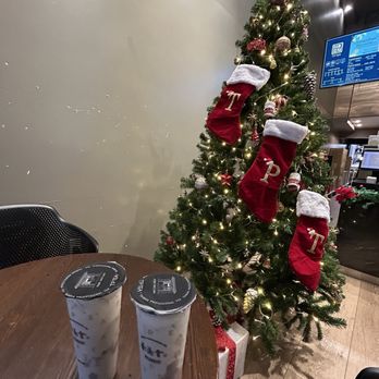 TP TEA - My friend and I got the same drink!   Taro milk tea - A Christmas tree decorated with ornaments, string lights, and three red stockings with white cuffs and gold lettering is positioned next to a wooden table. On the table, there are two cups of iced drinks with black lids, likely bubble tea or similar beverages. The setting appears to be an indoor cafe or restaurant, with a wall, a chair, and a digital menu screen visible in the background. - Coffee & Tea Near Me - Berkeley, California