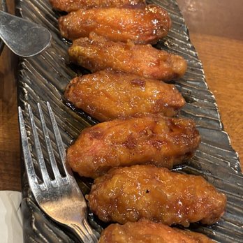 Colette - Cola Wings - Six glazed chicken wings are arranged on a black textured serving plate, with a fork and spoon nearby, on a wooden table. - Breakfast & Brunch Near Me - Pasadena, California