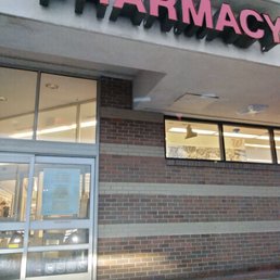 Store - The photo depicts the exterior of a Walgreens pharmacy store, featuring a large pink sign with the word 'PHARMACY' mounted above the entrance. The building has a brick wall with a section of glass windows and doors, allowing a view into the store interior. The image appears to be taken from inside a vehicle, with part of the dashboard visible at the bottom.