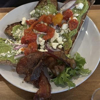 Jack Rabbit - A plate of avocado toast with two slices of toasted bread topped with mashed avocado, cherry tomatoes, crumbled cheese, and sliced red onions. The dish is garnished with additional cherry tomatoes and crumbled cheese, with a side of crispy bacon and fresh greens. The plate is placed on a wooden table, and part of a person's arm and watch are visible in the background. - Bars Near Me - Buffalo, New York