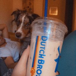 Sf iced sugar cookie breve with a soft top (good with white coffee too) - The image shows a person holding a large, clear plastic cup of iced coffee from Dutch Bros, with a blue straw and a label visible. In the background, a dog with a merle coat pattern looks toward the camera, and part of a person lying on a couch is also visible. The setting appears to be indoors, with warm lighting and home decor elements.
