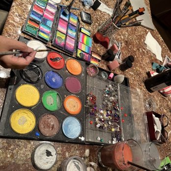 A Face Painter - A variety of face paints, brushes, and rhinestones are organized on a marble table, with makeup tools and supplies for face painting and cosmetics. - Face Painting Near Me