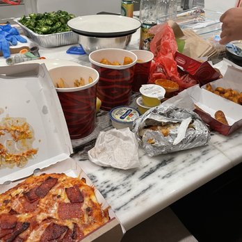 Sheetz - A table covered with food and drink items from a casual gathering, including an open pizza box with pepperoni pizza, snack boxes with fried foods, cups with beverages, and various food containers. The table surface is cluttered with wrappers, foil, and utensils, indicating an informal meal or party setting. - Convenience Stores Near Me - Hilliard, Ohio