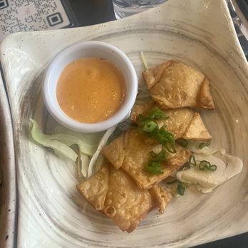 Bae Bae's Kitchen - Garlic Cheese Wontons - Fried wontons are arranged on a ceramic plate, garnished with chopped green onions. A small bowl of dipping sauce is placed on the plate, alongside some shredded cabbage. The setting includes a glass of water, a QR code, and utensils on a dark table surface, indicating a restaurant environment. - Korean Near Me - Pittsburgh, Pennsylvania