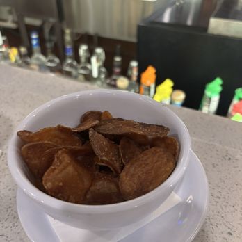 Barrel House - Homemade Chips - A white bowl filled with homemade potato chips is placed on a saucer with a napkin on a bar counter, with a blurred bar setting in the background. - American Near Me - Bloomington, Illinois
