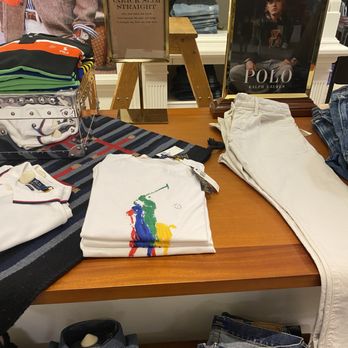 Dillard's - Polo Department - Clothing items such as white pants, a colorful polo shirt, and denim jeans are arranged on a wooden table in a retail store. - Department Stores Near Me - Saint Louis, Missouri