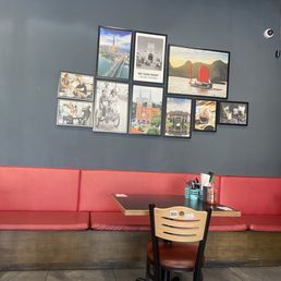Vietnam pics - The image shows a section of a restaurant interior featuring a red cushioned bench along a gray wall decorated with multiple framed photographs of cityscapes, landmarks, and people. A small wooden table with a black top is placed in front of the bench, with a chair positioned at the table. On the table, there are utensils, a small container, and a bottle. The floor is tiled with large gray tiles, and a security camera is visible on the wall.