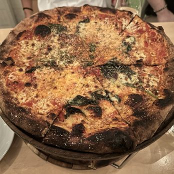 Gufo - I asked for my Margarita extra crispy. - A pizza with a thin, slightly charred crust is displayed on a wooden tray. The pizza is topped with melted cheese, tomato sauce, and fresh basil leaves. It appears to be freshly baked, with some areas of the crust showing signs of charring. The setting includes a table with glasses, a water bottle, and plates, indicating a dining environment. - Pizza Near Me - Cambridge, Massachusetts
