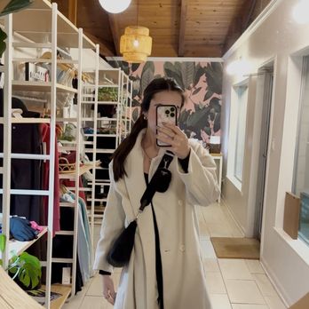 Organic Pilates - A woman is capturing a mirror selfie inside a clothing store, wearing a long beige coat and carrying a black shoulder bag. The store features wooden ceiling panels, hanging light fixtures, and shelves filled with clothing and accessories. The mirror has a round wooden frame, and the store has a bright, modern interior with natural light coming through the windows. - Pilates Near Me - Redding, California