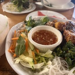 A plate of Vietnamese cuisine includes rice noodles, shredded lettuce, sliced cucumbers, shredded carrots, chopped green onions, and fresh herbs. A small bowl of dipping sauce with garlic and chili is placed in the center. The dish is served on a white plate, with additional plates and a bowl of soup visible in the background, indicating a restaurant setting.