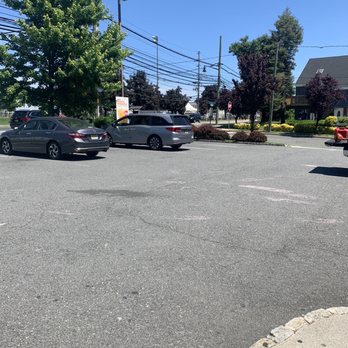 Dunkin Donuts - Parking - A parking lot with several parked cars, including sedans and minivans, is shown under a clear blue sky. There are trees and landscaped bushes along the perimeter, and power lines run across the background. A building with a sloped roof is visible on the right side, and the area appears to be part of a commercial or retail space. - Coffee & Tea Near Me - Secaucus, New Jersey