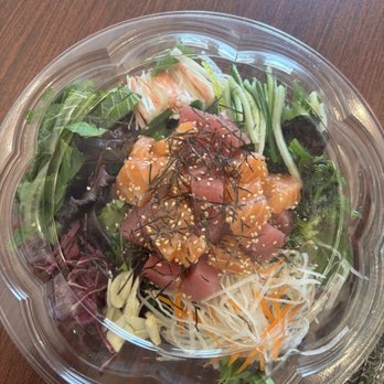 K-Town Takeout - A round, clear plastic container contains a mixed salad with various ingredients including shredded lettuce, carrots, seaweed, and diced tuna topped with sesame seeds and shredded nori. The salad is arranged with different colors and textures, and the container is placed on a wooden table surface. - Korean Near Me - Frederick, Maryland