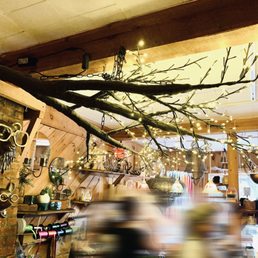 Glow Flow - A branch decorated with string lights is suspended from the ceiling in a candle shop, with blurred figures below and shelves of candles and decor.