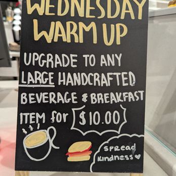 Vons - My new favorite!!!!! - A black chalkboard sign with yellow and white handwritten text displays a Wednesday warm-up promotion. It offers an upgrade to any large handcrafted beverage and breakfast for $10. The sign includes illustrations of a coffee cup and a sandwich, along with the message 'spread kindness' and a heart symbol. The sign is positioned on a small wooden stand on a counter, with a blurred background of a cafe or coffee shop interior. - Grocery Near Me - Camarillo, California