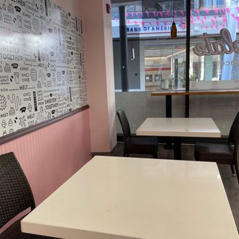 Chocolate Bash - a table and chairs in a restaurant - The image shows the interior of a dessert shop with two white tables and black woven chairs. One table is positioned near a pink wall with decorative black and white graphic art related to chocolates and sweets. Large glass windows reveal a view of the street outside, including a Bank of America branch and pink bunting. The space has a modern, clean design suitable for casual dining or dessert consumption. - Desserts Near Me - Phoenix, Arizona