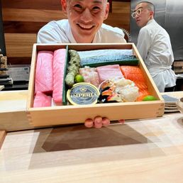 Chef Okano and the sushi selection for the evening - A man with a shaved head and white shirt is smiling and holding a wooden box filled with various types of sushi, including sashimi, nigiri, and rolls. The setting appears to be a restaurant with wooden and metallic interior elements, and another person in the background is wearing glasses and a white chef's coat, facing away.
