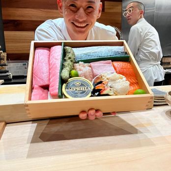 Hiden - Chef Okano and the sushi selection for the evening - A man with a shaved head and white shirt is smiling and holding a wooden box filled with various types of sushi, including sashimi, nigiri, and rolls. The setting appears to be a restaurant with wooden and metallic interior elements, and another person in the background is wearing glasses and a white chef's coat, facing away. - Japanese Near Me - Miami, Florida