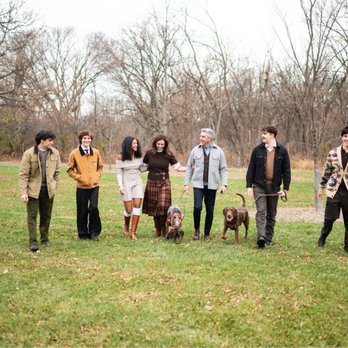 Ben Lipowitz Photo - A group of seven individuals, including men and women, walk together in a park with leafless trees and a grassy field. Two dogs are with them, one on a leash held by a man and the other walking freely. The people are dressed in casual fall attire, and the scene appears to be during late autumn or early winter. - Session Photography Near Me