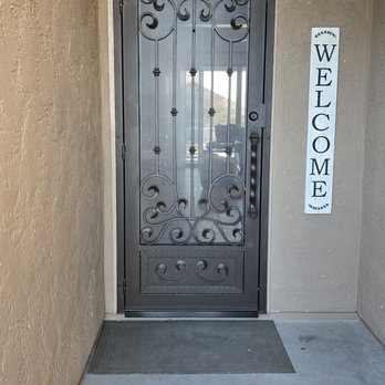 First Impression Ironworks - Front security door - A metal security door featuring decorative scrollwork is set into a beige stucco wall. The door has a handle on the right side and a small lock. To the right of the door, a vertical "Welcome" sign is mounted on the wall. The ground in front of the door is concrete with a small doormat placed directly in front of the door. - Fences & Gates Near Me - Gilbert, Arizona