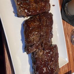 Three beef ribs with a glossy barbecue sauce coating are arranged on a white rectangular plate. The ribs are placed on a wooden table, with part of a drink and a fork visible nearby. The image captures the presentation of the ribs, highlighting the sauce and the meat's texture.