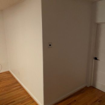 Knotty's Custom Carpentry - Den. - The image depicts a corner of an interior room with a white painted wall and a wooden floor. A light switch is mounted on the wall near the corner. To the right, there is a door with a black handle. The ceiling appears textured, and the lighting is dim, casting shadows on the wall. - Handyman Near Me