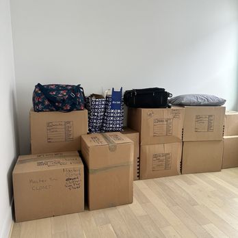 Optimus Moving Services - Boxes labelled for the closet dumped in a room that's definitely NOT the closet. - Several cardboard moving boxes are stacked against a wall, with bags and a pillow placed on top, in a room with light-colored wooden flooring. - Movers Near Me - Katy, Texas