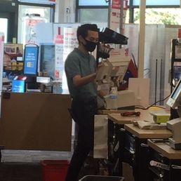 Derek working diligently - A man wearing a black face mask and glasses is working at a checkout counter in a store, scanning items and handling a cash register.