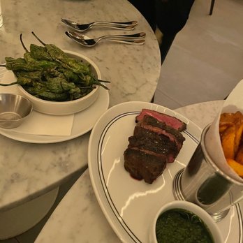 Vela - Shishito peppers, 8oz steak - A marble table holds a plate with sliced steak, a bowl of green sauce, and a dish of roasted peppers, with utensils nearby. - Venues & Event Spaces Near Me - Toronto, Ontario
