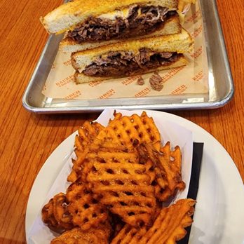Backporch Drafthouse - Temple - Brisket sandwich and sweet potato fries - The image shows a meal served on a wooden table, including a metal tray with a brisket sandwich cut in half, held together with a skewer, and accompanied by French fries. In front, there is a white plate with waffle fries and pieces of fried chicken. The setting appears to be a casual dining environment, possibly a restaurant or bar, with various items like napkins, utensils, and a drink visible in the background. - American Near Me - Temple, Texas