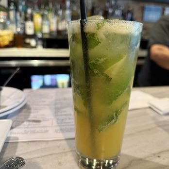Cafe con Leche - Passion fruit mojito - A tall glass contains a passion fruit mojito with lime slices, mint leaves, and a black straw, placed on a bar counter with a blurred bar background. - Mexican Near Me - Chicago, Illinois