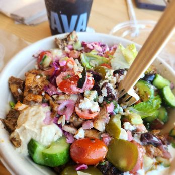 CAVA - Build your own bowl of deliciousness - A salad bowl contains cherry tomatoes, cucumbers, pickles, crumbled cheese, and other vegetables, with a fork inserted. A drink in a plastic cup with a straw is visible behind. - Mediterranean Near Me - Dedham, Massachusetts