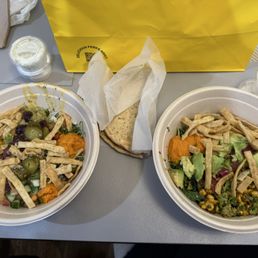 Greens + Grains Bowl - Two salad bowls with various vegetables, toppings, and shredded chicken, along with a pita bread, are placed on a gray table.