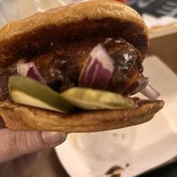 Rib Sandwich - A close-up image shows a barbecue sandwich with a toasted bun, filled with pulled meat, pickles, and sliced onions. The sandwich is held in a person's hand, with a blurred background that includes a drink and some indistinct objects, suggesting a casual dining setting.