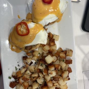 Perk Up Kitchen - Eggs Benedict - Two servings of eggs Benedict topped with hollandaise sauce and sliced chili peppers are presented on a white rectangular plate. The eggs are poached with the sauce drizzled over them. A side of diced breakfast potatoes, seasoned and garnished with herbs, is placed alongside. The dish is served on a white table with a fork visible at the edge of the plate. - Breakfast & Brunch Near Me - Bloomington, Minnesota