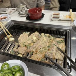 Pork Belly - Slices of pork belly are cooking on a built-in grill at a restaurant table, with garlic and green chili peppers nearby. The table has plates with sauces, chopsticks, and other dining utensils, indicating a Korean BBQ dining experience.