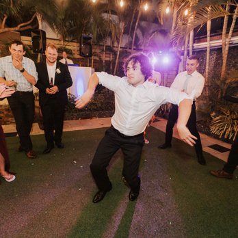 Vision DJs - Our guests enjoying the DJ's song selections - A group of guests is dancing and celebrating outdoors, with some clapping and smiling. The scene includes a DJ with equipment and lighting effects, and the area is decorated with string lights and tropical plants. The event appears lively and festive, with people dressed in semi-formal attire. - DJs Near Me - Miami, Florida