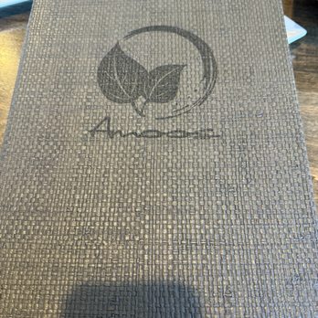 Amoo's Restaurant - A menu cover with a woven, textured surface displays a logo of three leaves within a circle and the text 'Amoo's' underneath. The menu is placed on a wooden table, with utensils and a plate partially visible around it. The cover appears to be made of a durable material suitable for a restaurant setting. - Persian/Iranian Near Me - McLean, Virginia