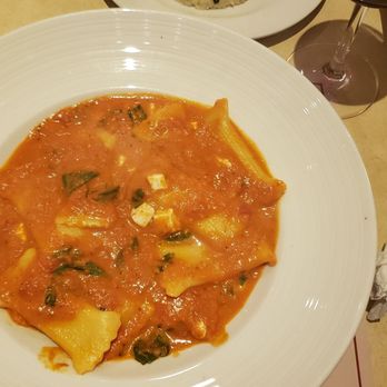 La Piazza - Perfect pasta pillows - A white round plate holds pasta with a tomato-based sauce, garnished with small pieces of cheese and herbs. The plate is placed on a dining table with utensils, a crumpled napkin, and a basket of bread nearby. The setting suggests a casual Italian restaurant meal. - Pizza Near Me - Plainview, New York