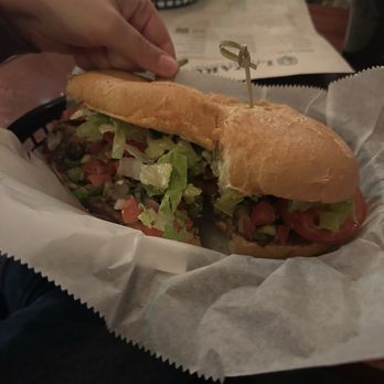 Lazarus Brewing - Torta - A large sandwich with lettuce, tomato, and meat is placed on white paper in a restaurant. A hand is holding the sandwich, and a menu and drink are visible in the background. - Breweries Near Me - Austin, Texas