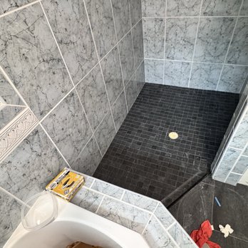 4 Ace Tile - A shower area features gray marble tiles on the walls and a black tiled floor with a drain in the center. Toiletries such as bottles and a soap dish are placed on a wall-mounted shelf. The image also shows part of a bathtub with a brown paper bag and a box of disposable razors nearby, along with some scattered items on the floor. - Tiling Near Me - Modesto, California