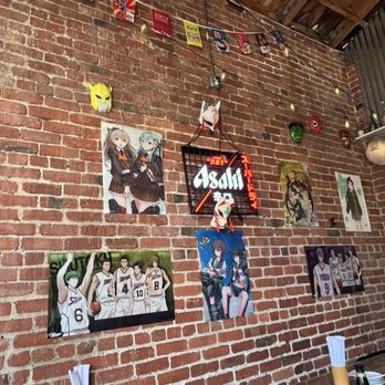 Kinda Izakaya - Other wall decor - The image shows a brick wall decorated with various anime posters, masks, and a neon Asahi sign, indicating a Japanese-style restaurant or bar. The wall features posters of anime characters, masks resembling superhero and traditional styles, and a string of small decorative lights. The setting includes wooden tables with chopsticks and condiments, suggesting a dining environment with Japanese cultural decor. - Tapas/Small Plates Near Me - Berkeley, California