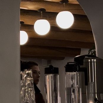 La Reina - A woman is seen behind a bar counter with glassware and beverage dispensers, with hanging spherical lights and wooden ceiling panels in the background. - Bars Near Me - Santa Fe, New Mexico