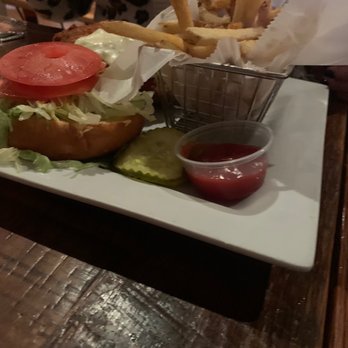Public Kitchen - Chicken Sandwich - A plate displays a burger with sliced tomato, lettuce, and pickles, served with a side of French fries in a metal basket lined with paper. A small cup of ketchup is placed on the plate, and the meal is set on a wooden table. The background shows people in a casual dining environment. - Gastropubs Near Me - Wakefield, Massachusetts