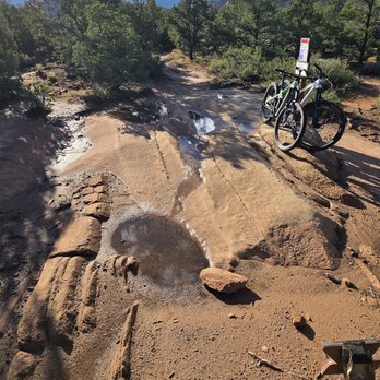 Hazard County Shuttle - A mountain biking trail features uneven, rocky terrain with small pools of water and scattered rocks. Two bikes are parked on the side of the trail, with one leaning against a signpost. The background includes trees, distant mountains, and a partly cloudy sky, indicating an outdoor, natural setting suitable for mountain biking activities. - Transportation Near Me