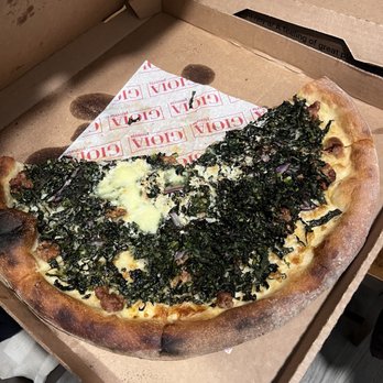 Gioia Pizzeria - Kale sausage - A partially eaten pizza with a crust, topped with chopped kale, melted cheese, and meat, inside a cardboard pizza box. The box has branding and is placed on a wooden surface. The pizza appears to be a specialty with vegetable and meat toppings. - Pizza Near Me - Berkeley, California