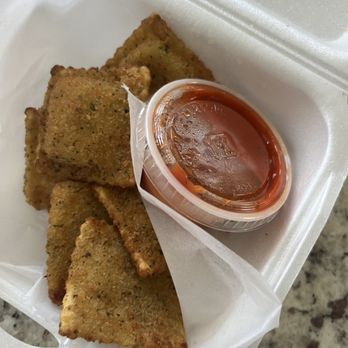 Pizza World - Ravioli - Four fried ravioli pieces are arranged in a foam takeout container, lined with white paper. A small plastic cup of marinara sauce is placed on top of the ravioli, with the sauce appearing glossy and red. The container is on a speckled countertop, and the image captures a close-up view of the appetizer. - Pizza Near Me - Branson, Missouri