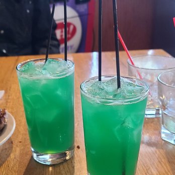 Dunaways - two drinks on a table - Two tall glasses filled with bright green liquid labeled "Liquid Marijuana" sit on a wooden table, with ice and straws, in a restaurant setting. - Pubs Near Me - Troy, Ohio
